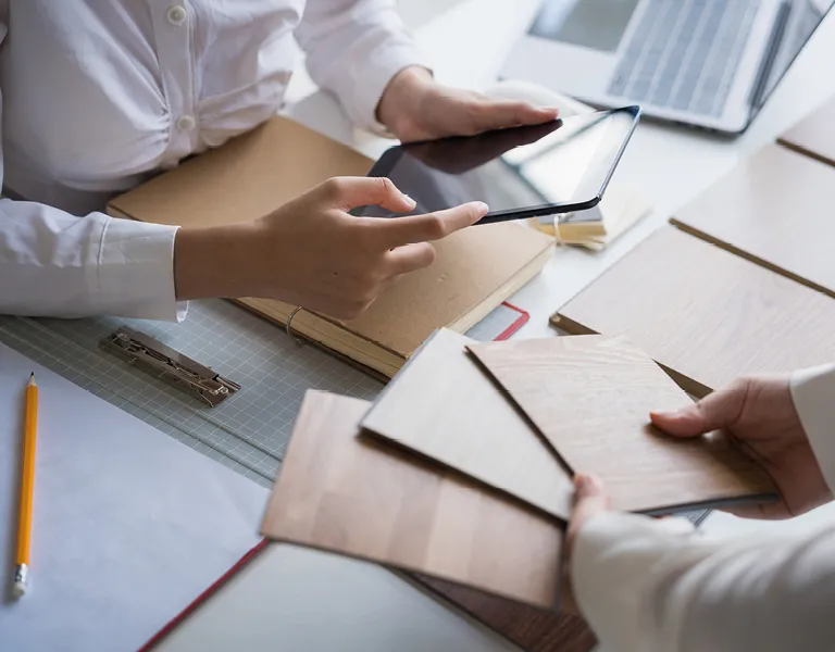 Zwei Personen vergleichen Holzdekore für Küchenmöbel auf einem Tisch, während ein Tablet zur Visualisierung genutzt wird, daneben liegen Unterlagen, ein Laptop und ein Bleistift.